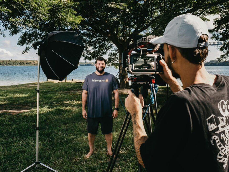 Filmmaker with video camera on tripod shooting video of smiling man standing on glade on coast of pond
